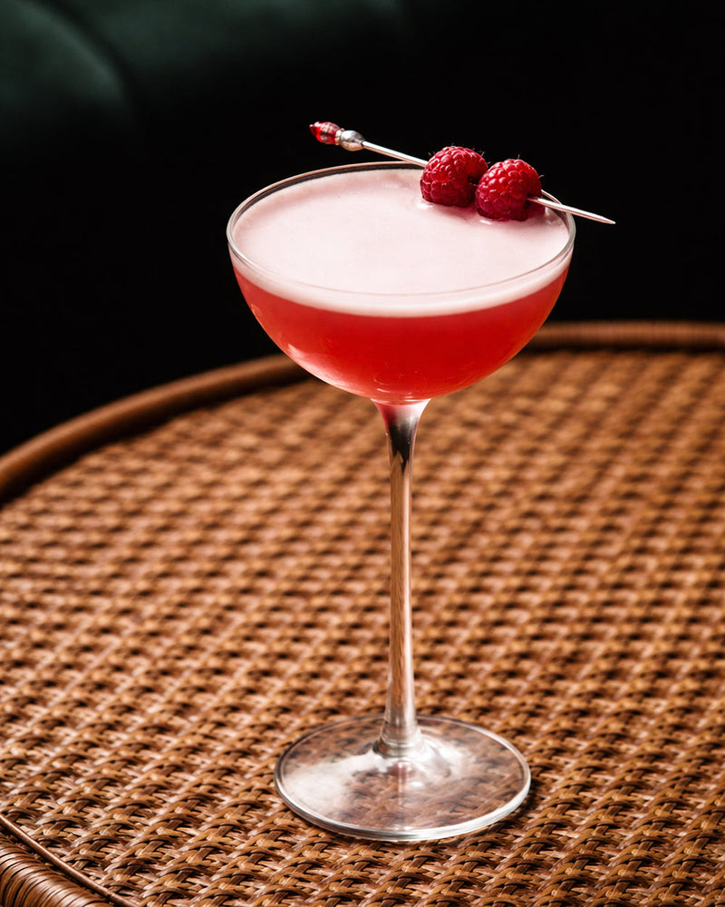 a pink cocktail with two raspberries as garnish sitting on a rattan tabletop